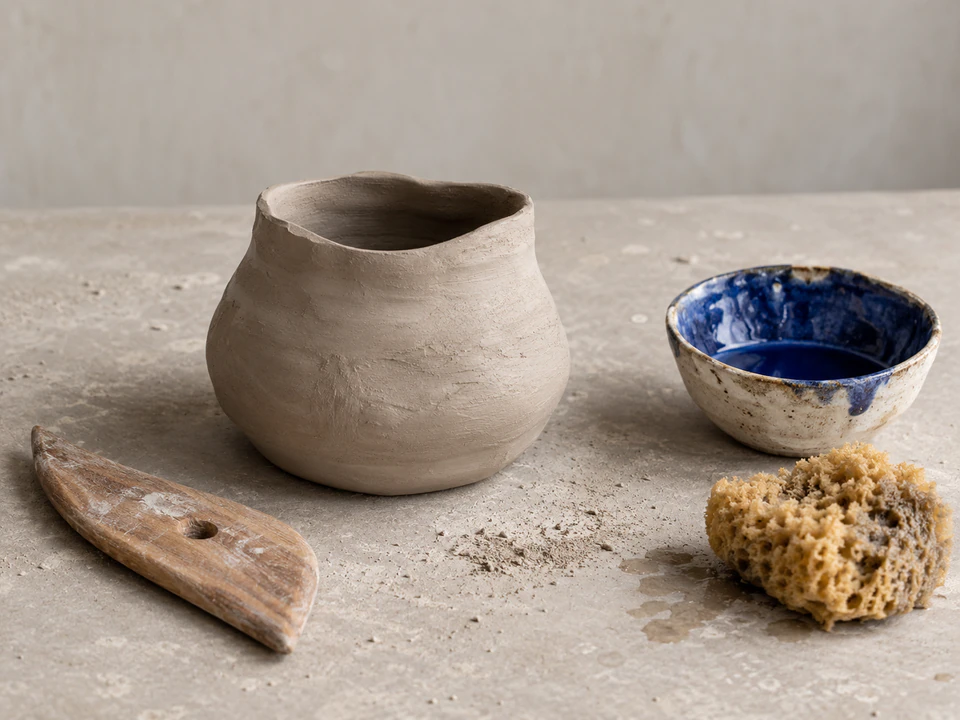 An unglazed clay vessel, a bowl of blue glaze and pottery tools on a studio workbench