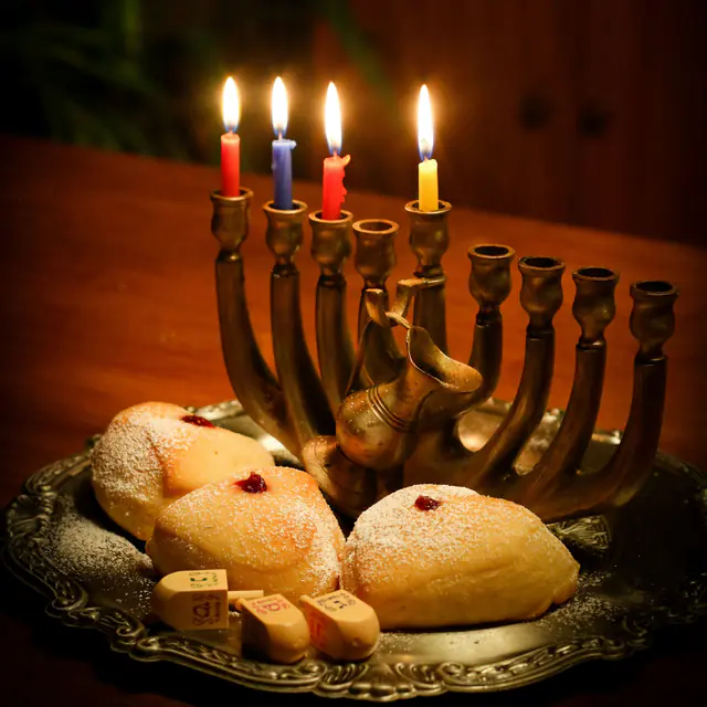 A brass menorah with lit candles beside a plate of sufganiyot and wooden dreidels