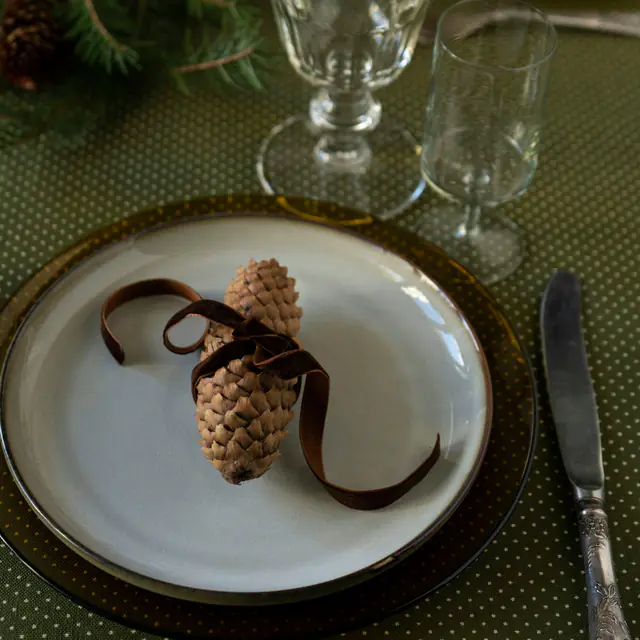 A holiday place setting: a pine cone tied with brown ribbon on a plate, glasses and fir sprigs