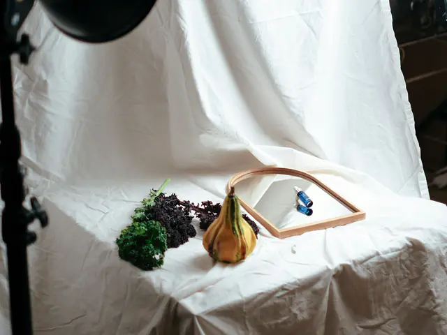 A small home photo setup: a studio light and softbox aimed at a gourd, kale and a wooden frame on white linen