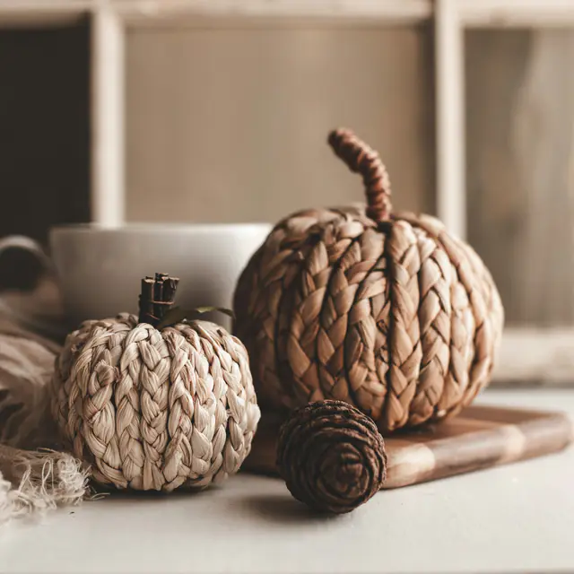 Two hand-braided pumpkins with twig stems on a wooden board beside a white mug and a linen throw