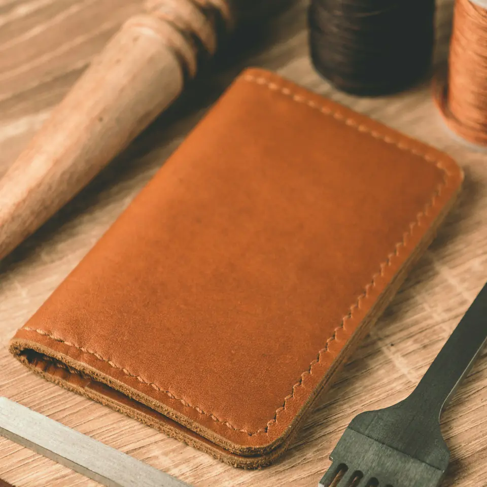 A hand-stitched tan leather card wallet on a workbench beside leatherworking tools and spools of thread