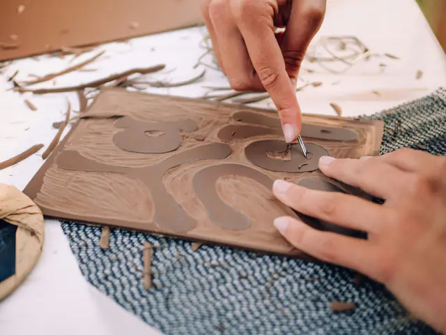 Hands carving a curving pattern into a linocut block, with lino shavings scattered across the table
