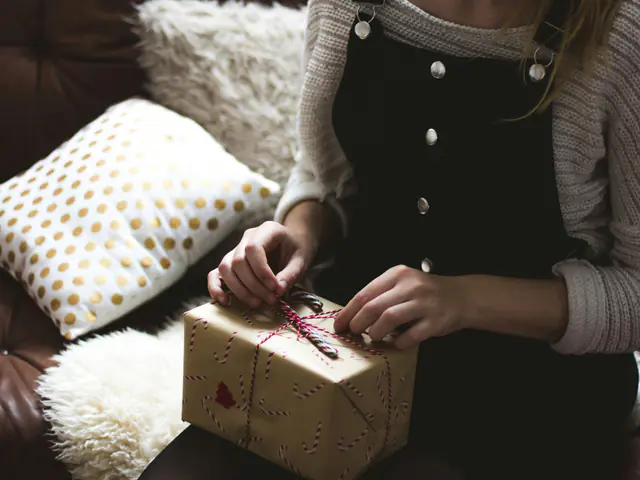 A person on a sofa untying the red-and-white string on a parcel wrapped in printed brown paper