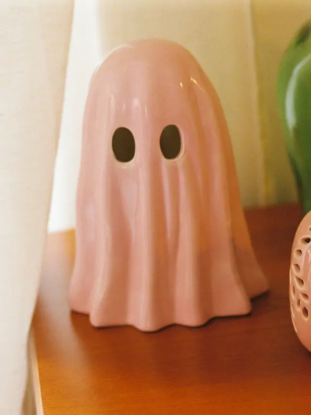A pastel pink ceramic ghost with cut-out eyes on a wooden table in warm window light