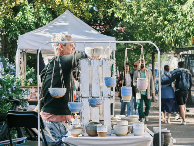 A potter at his outdoor market booth with hanging planters and stacks of glazed bowls on a white rack