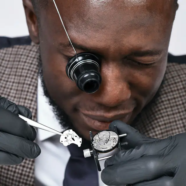 A watchmaker in Accra wearing a loupe, fitting a part into a watch movement with gloved hands