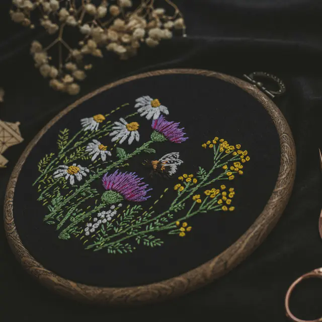 Hand embroidery of wildflowers and a bee in a wooden hoop on black linen, beside stork scissors