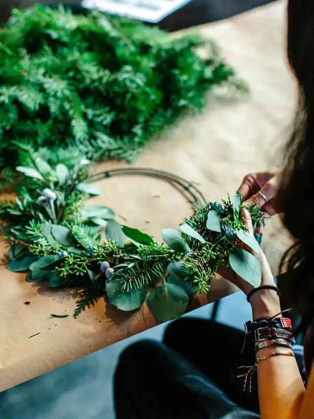 Hands shaping a fresh green wreath of fir and eucalyptus on a kraft-paper covered table